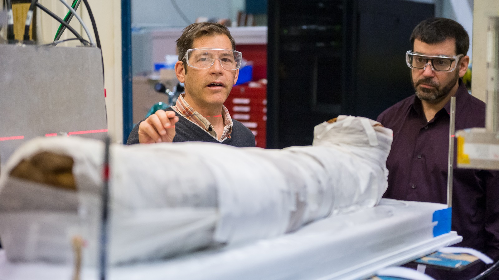 Argonne physicist and group leader Jonathan Almer, left, talks with Dan Silverstein of the Block Museum of Art at Northwestern University in 2017. Almer points out the red laser beams that will guide the X-ray probe of the Egyptian mummy in the foreground. (Image by Mark Lopez / Argonne National Laboratory.)