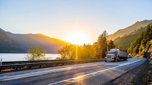 Semi-truck on highway, mountains, water, sun in background. (Image by Shutterstock/Vitpho.)