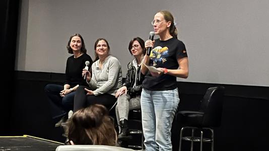 Four female scientists sit in front of a movie screen and speak into microphones.