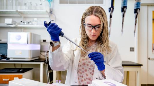 A scientist in a lab coat and protective eye wear transfers a sample from a pipette to a test tube.