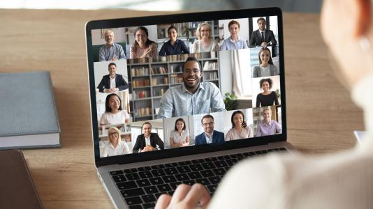 An over the shoulder image of person sitting at table looking at an online meeting (represented by thumbnail images of different people on the screen) on a laptop.