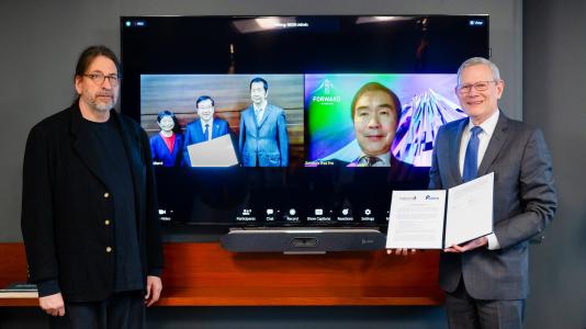 Two men are standing next to a video screen that shows four people from Japan who participated in a virtual signing event.