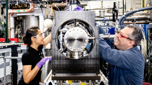 Two scientists smile as they adjust large metal scientific equipment