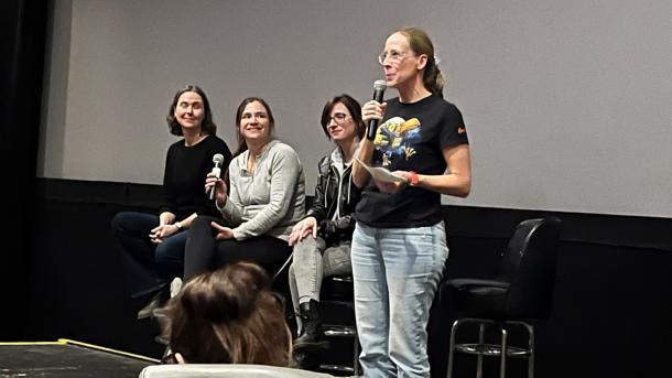 Four female scientists sit in front of a movie screen and speak into microphones.